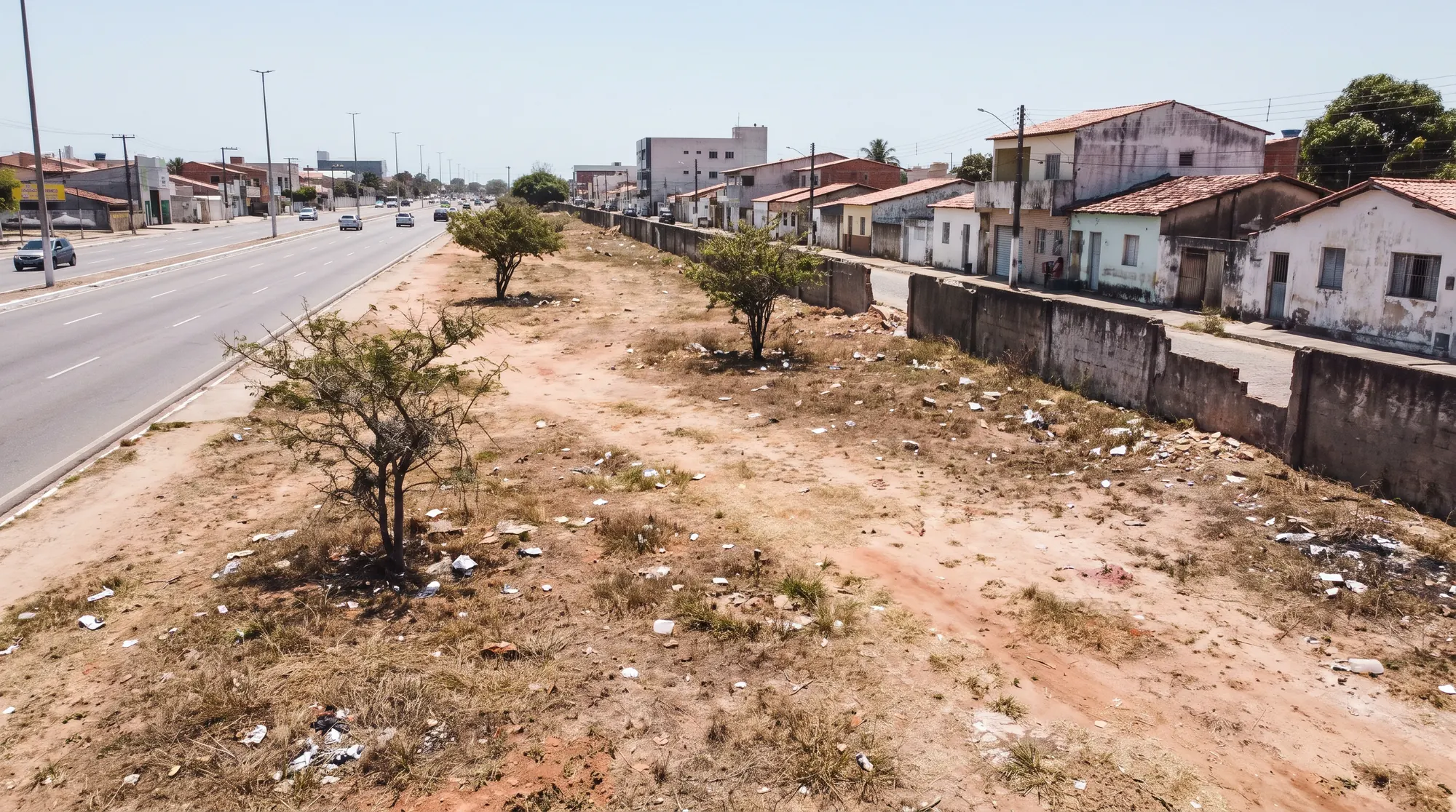 Faixa de terreno abandonada ao lado de uma avenida, com terra seca e poucas árvores.